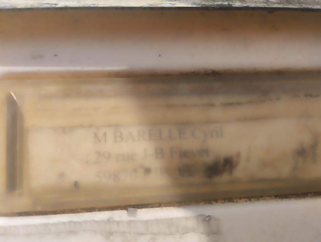 Boîte aux lettres floue derrière une vitre sale, coordonnées partiellement illisibles.